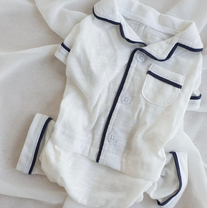 May include: White pajamas with a navy blue trim and buttons. The pajamas have a pocket on the left side.