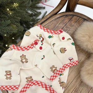 May include: A white baby onesie with a red and white gingham trim. The onesie has a Christmas theme with bears, trees, and presents.