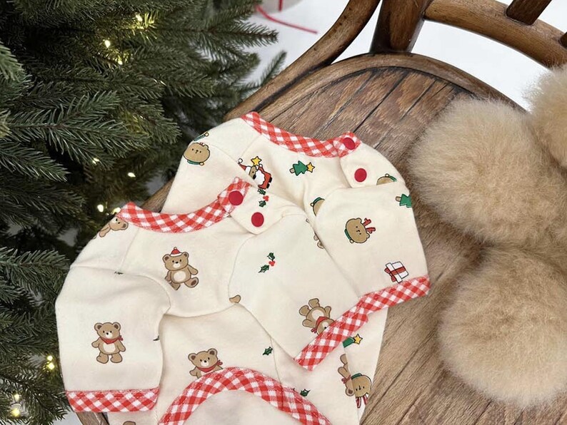 May include: A white baby onesie with a red and white gingham trim. The onesie has a Christmas theme with bears, trees, and presents.
