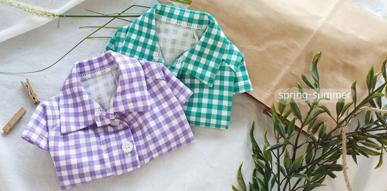 May include: Two checkered shirts, one purple and one green, with short sleeves and button closures. The shirts are laid out on a white surface with a brown paper bag and green foliage in the background.