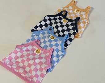 May include: Four checkered tank tops with a smiley face patch. The tank tops are in pink, blue, black, and yellow.