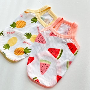 May include: Two sleeveless dog shirts with a summer fruit theme. One shirt features a pineapple pattern on a white background with a yellow trim. The other shirt features a watermelon pattern on a white background with a pink trim.