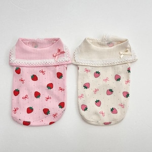 Puede incluir: Dos camisetas para mascotas, una rosa y otra crema, con un estampado de fresas. Las camisetas tienen una textura acanalada, un cuello de encaje y un pequeño lazo. La camiseta rosa tiene fresas rojas, mientras que la crema tiene fresas rosas.