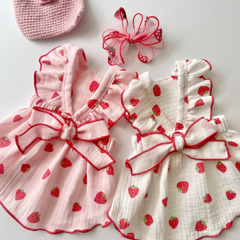 May include: Two pink and white sleeveless dresses with strawberry print and red ribbon bows. The dresses have a ruffled neckline and are made of a soft, lightweight fabric.