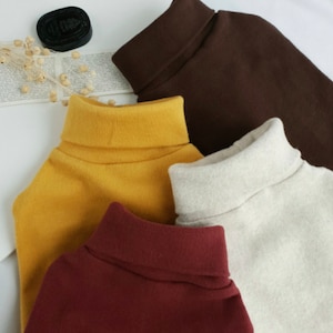 May include: Four turtleneck sweaters in different colors: brown, yellow, beige, and burgundy. The sweaters are folded and stacked on top of each other.