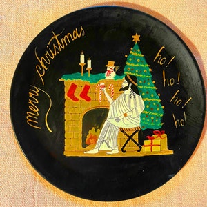 Op de afbeelding: Een zwarte keramische plaat met een handgeschilderd kersttafereel. De scène toont een open haard met kousen, een sneeuwpop, een kerstboom en een persoon die een glas wijn vasthoudt. De tekst "Merry Christmas" en "Ho! Ho! Ho!" staat op de plaat.