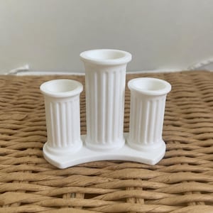 May include: A white candle holder featuring three fluted columns of varying heights arranged on a curved base. The central column is taller than the two flanking columns, and each column has a circular opening at the top for holding a candle.