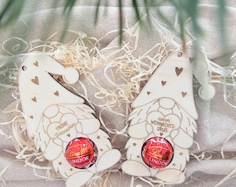 May include: Two light-colored wooden gnome ornaments with heart cutouts, each holding a round red Lindt Lindor chocolate truffle. The left gnome is engraved with "Frohe Weihnachten" and the right gnome with "kleiner WEIHNACHTS-GRUB".