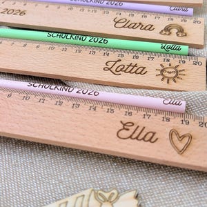 May include: Wooden rulers with engraved names and decorative elements, alongside colored pencils. Each ruler is approximately 8 inches long and features a name, a heart, a sun, or a rainbow. The pencils are light blue, purple, and green.