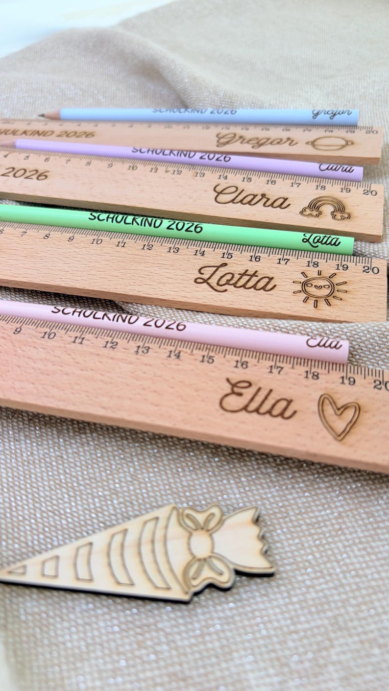 May include: Wooden rulers with engraved names and decorative elements, alongside colored pencils. Each ruler is approximately 8 inches long and features a name, a heart, a sun, or a rainbow. The pencils are light blue, purple, and green.
