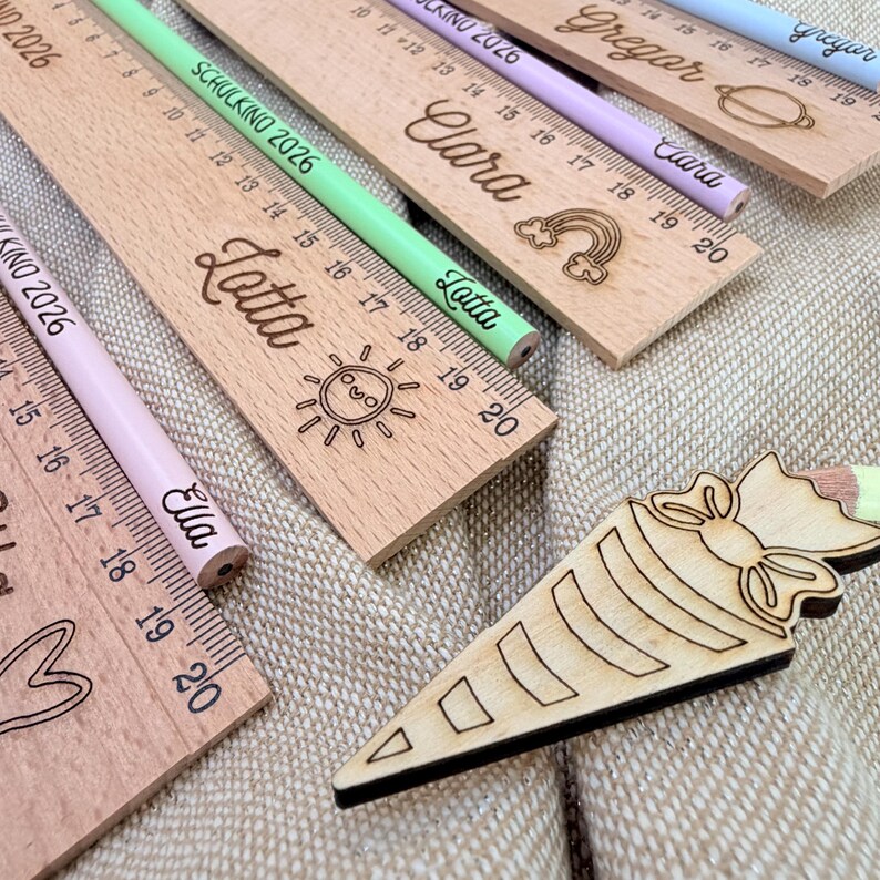 May include: Wooden rulers and pencils with engraved names and designs. The rulers have centimeter markings and the pencils are in various colors. The image includes the names "Clara", "Lotta", "Ella", and "Gregor".