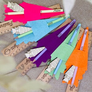 May include: A collection of colorful felt pencil holders in pink, blue, purple, green, and orange, each with a pencil and a wooden ruler. The pencils are labeled with names and the year 2025. The rulers have names and measurements.