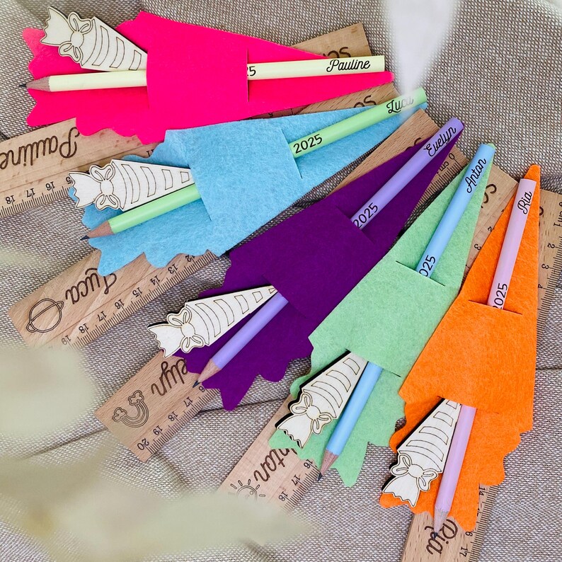May include: A collection of colorful felt pencil holders in pink, blue, purple, green, and orange, each with a pencil and a wooden ruler. The pencils are labeled with names and the year 2025. The rulers have names and measurements.
