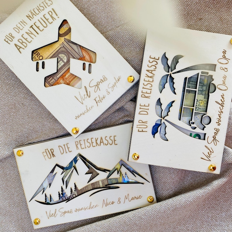 Puede incluir: Tres cajas de madera para dinero con dise&ntilde;os recortados. La primera caja tiene un avi&oacute;n con el texto "F&Uuml;R DEIN N&Auml;CHSTE ABENTEUER!" La segunda caja tiene monta&ntilde;as con el texto "F&Uuml;R DIE REISEKASSE Viel Spa&szlig; w&uuml;nschen Nico & Marie". La tercera caja tiene palmeras y una camioneta con el texto "F&Uuml;R DIE REISEKASSE Viel Spa&szlig; w&uuml;nschen Oma & Opa".