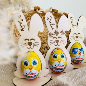 Puede incluir: Tres soportes para huevos de Pascua de madera con forma de conejo con los nombres "Mats", "Julie" y "Paul" escritos en ellos. Cada conejo sostiene un huevo Kinder Surprise amarillo.