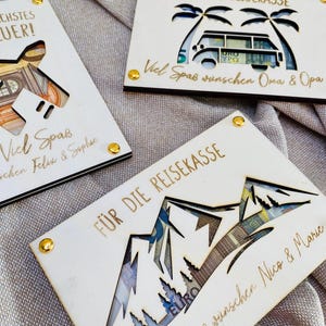 Puede incluir: Tres cajas de madera para dinero con dise&ntilde;os recortados. La primera caja tiene un dise&ntilde;o de avi&oacute;n y el texto "Liebstes Quer!" La segunda caja tiene un dise&ntilde;o de palmera y autocaravana con el texto "F&uuml;r die Reisekasse Viel Spa&szlig; w&uuml;nschen Oma & Opa". La tercera caja tiene un dise&ntilde;o de monta&ntilde;a con el texto "F&uuml;r die Reisekasse Viel Spa&szlig; w&uuml;nschen Nico & Marie".