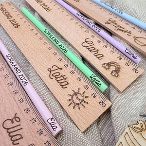 May include: Wooden rulers with engraved names and measurements in inches. Each ruler has a matching colored pencil. The rulers are personalized with names like "Lotta" and "Clara", and feature decorative elements like a sun and rainbow.