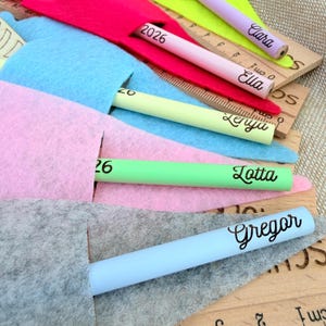 May include: A collection of colorful pencils, each with a name printed on it, are partially inserted into felt pouches. The pencils are in various colors, including pink, green, and blue. Wooden rulers with centimeter markings are also visible.