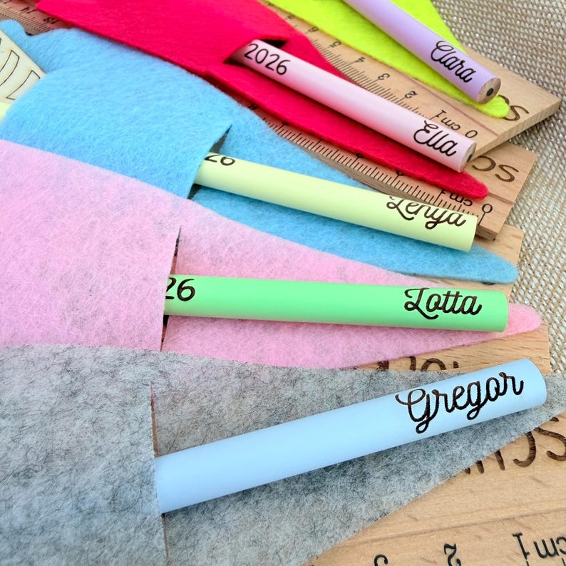 May include: A collection of colorful pencils, each with a name printed on it, are partially inserted into felt pouches. The pencils are in various colors, including pink, green, and blue. Wooden rulers with centimeter markings are also visible.