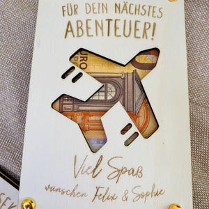 Puede incluir: Una tarjeta de madera con un avi&oacute;n recortado. La tarjeta tiene el texto "F&uuml;r dein n&auml;chstes ABENTEUER!" y "Viel Spa&szlig; w&uuml;nschen Felix & Sophie".