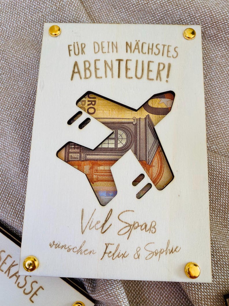 Puede incluir: Una tarjeta de madera con un avi&oacute;n recortado. La tarjeta tiene el texto "F&uuml;r dein n&auml;chstes ABENTEUER!" y "Viel Spa&szlig; w&uuml;nschen Felix & Sophie".