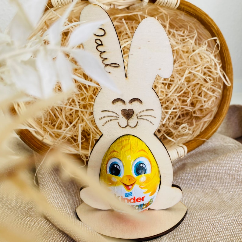 Puede incluir: Corte de conejo de Pascua de madera con el nombre "Paul" grabado en el lateral. El conejo sostiene un huevo de chocolate amarillo con un dise&ntilde;o de pollito.