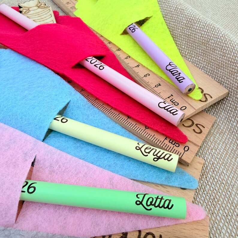 May include: A collection of colorful pencils with names printed on them, resting on wooden rulers and nestled in felt pouches. The pencils are in shades of pink, green, purple, and yellow. The rulers have centimeter markings.