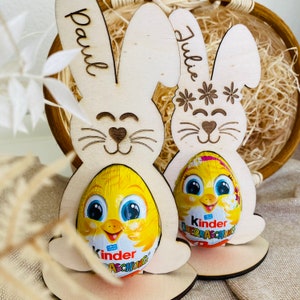 Puede incluir: Dos soportes de huevos de Pascua de conejo de madera con los nombres "Paul" y "Julie" escritos en ellos. Cada conejo sostiene un huevo de chocolate Kinder Surprise.