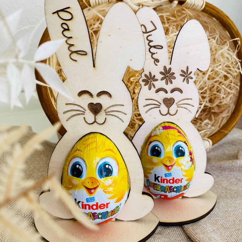 Puede incluir: Dos soportes de huevos de Pascua de conejo de madera con los nombres "Paul" y "Julie" escritos en ellos. Cada conejo sostiene un huevo de chocolate Kinder Surprise.