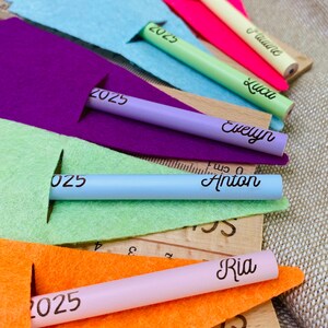 May include: A collection of colorful pencils, each with a name and the number 2025 printed on them. The pencils are in various colors, including pink, green, and blue, and are placed on top of felt and wooden rulers.