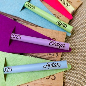 May include: A collection of personalized pencils in various colors, including green, purple, and blue, each with a name and the year 2025 or 025 printed on them. The pencils are displayed with wooden rulers and colorful felt cutouts.