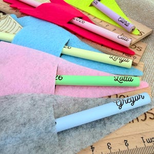 May include: A collection of colorful pencils with personalized names, each nestled in a felt sleeve. The pencils are in various colors, including pink, green, and blue. Wooden rulers with centimeter markings are also visible.