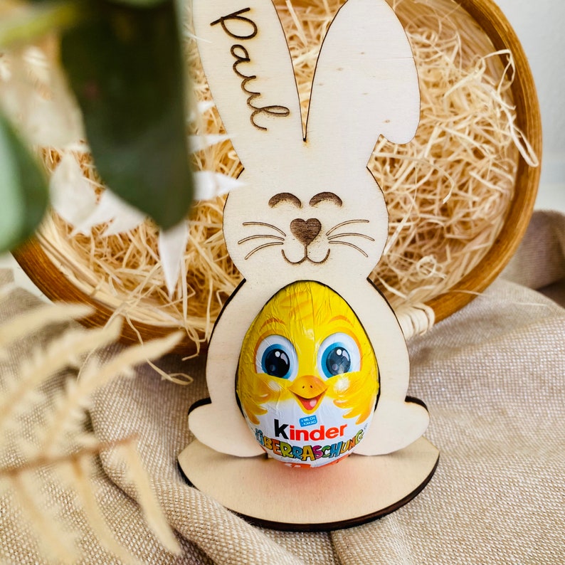 Puede incluir: Porta huevos de Pascua de madera en forma de conejo con el nombre "Paul" escrito en &eacute;l. El soporte sostiene un huevo Kinder Surprise amarillo con un dise&ntilde;o de pollito.