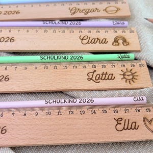 May include: Wooden rulers and pencils with names and the text "SCHULKIND 2026". The rulers are engraved with names and decorative elements. The pencils are pastel colors: blue, purple, green, and pink. The rulers measure up to 20 inches.