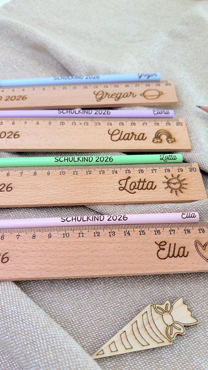May include: Wooden rulers and pencils with names and the text "SCHULKIND 2026". The rulers are engraved with names and decorative elements. The pencils are pastel colors: blue, purple, green, and pink. The rulers measure up to 20 inches.