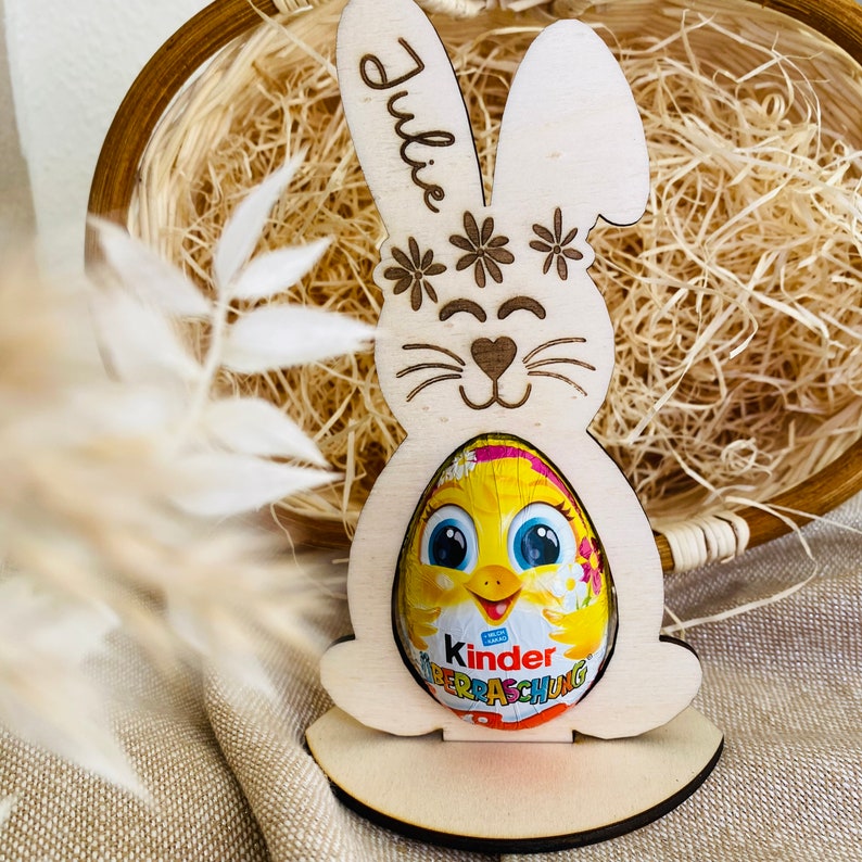 Puede incluir: Un recorte de conejo de madera con el nombre "Julie" escrito en &eacute;l. El conejo sostiene un huevo Kinder Surprise amarillo y rosa. El conejo est&aacute; de pie sobre una base de madera.