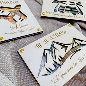 Puede incluir: Tres cajas de dinero de madera con recortes de un avi&oacute;n, un coche y monta&ntilde;as. Cada caja tiene un mensaje deseando al destinatario un feliz viaje e incluye el texto "Para la caja de viaje".