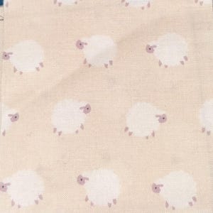 May include: Beige fabric with a repeating pattern of white sheep with purple accents. The sheep are stylized with rounded bodies and small legs, creating a whimsical design suitable for various craft projects.