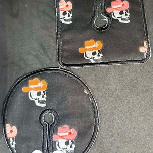 May include: Two black fabric nipple covers with a skull and cowboy hat design. The covers have a slit in the middle and two white buttons on the bottom.