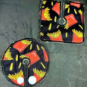 May include: Two black fabric patches with a red and yellow french fry design. The patches have a hole in the center and are meant to be sewn onto clothing.