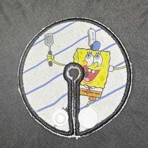 May include: A circular patch featuring the cartoon character SpongeBob SquarePants holding a spatula. The patch has a white background with blue stripes and a black border. The patch also has a black and white clasp.