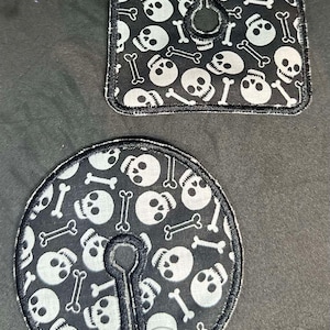 May include: Two black fabric nipple covers with a white skull and crossbones pattern. The covers have a hole in the center and are secured with a snap closure.