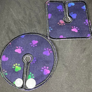 May include: Two dark blue fabric patches with a paw print pattern and a hole in the center. The patches have a black border and are sewn with white thread. The larger patch has two white buttons.
