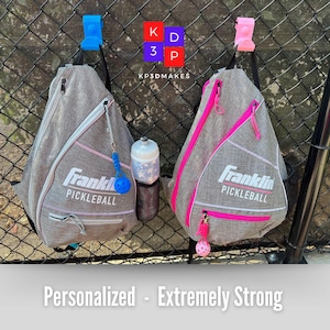 Op de afbeelding: Twee grijze Franklin Pickleball slingtassen met roze en witte accenten hangen aan een hek van gaas. Elke tas heeft een rits en een kleine bedel. Een waterfles en een blauwe en roze pickleball zijn ook zichtbaar. De tekst "Personalized - Extremely Strong" staat onderaan.