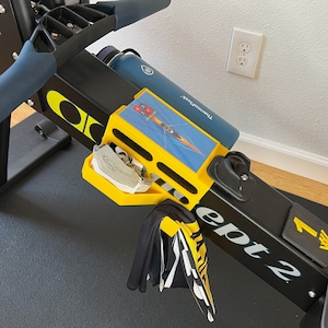 May include: A black rowing machine with a yellow and black accessory holder. The holder has a small water bottle, a white and gray object, and a black and white towel hanging from it. The machine has a black and white label that says "ept 2" and a yellow label that says "1 min".