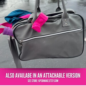 Op de afbeelding: Een grijze tas met lederlook, witte rits en handvatten. Een felroze accessoire met de naam "LAYLA" is aan het handvat bevestigd. In de tas zitten kleurrijke items, waaronder een paars en roze verpakt item en een blauw kledingstuk.