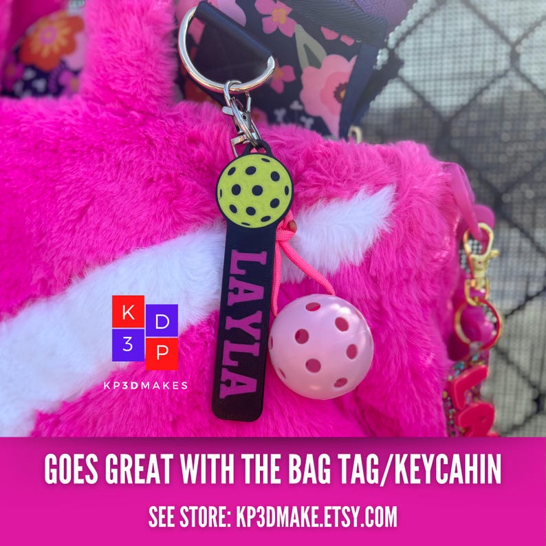 Op de afbeelding: Een roze pickleball sleutelhanger met een zwart label met de naam "LAYLA" in roze letters. De sleutelhanger heeft een gele pickleball met zwarte stippen en een roze pickleball met roze stippen. De achtergrond is een roze fuzzy tas.