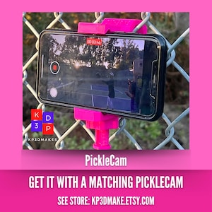 Op de afbeelding: Een roze PickleCam-telefoonhouder bevestigd aan een hek van gaas, met een smartphone die een pickleball-wedstrijd opneemt. De tekst "PickleCam" wordt onder de telefoon weergegeven. De merknaam "KP3DMAKES" is ook zichtbaar.