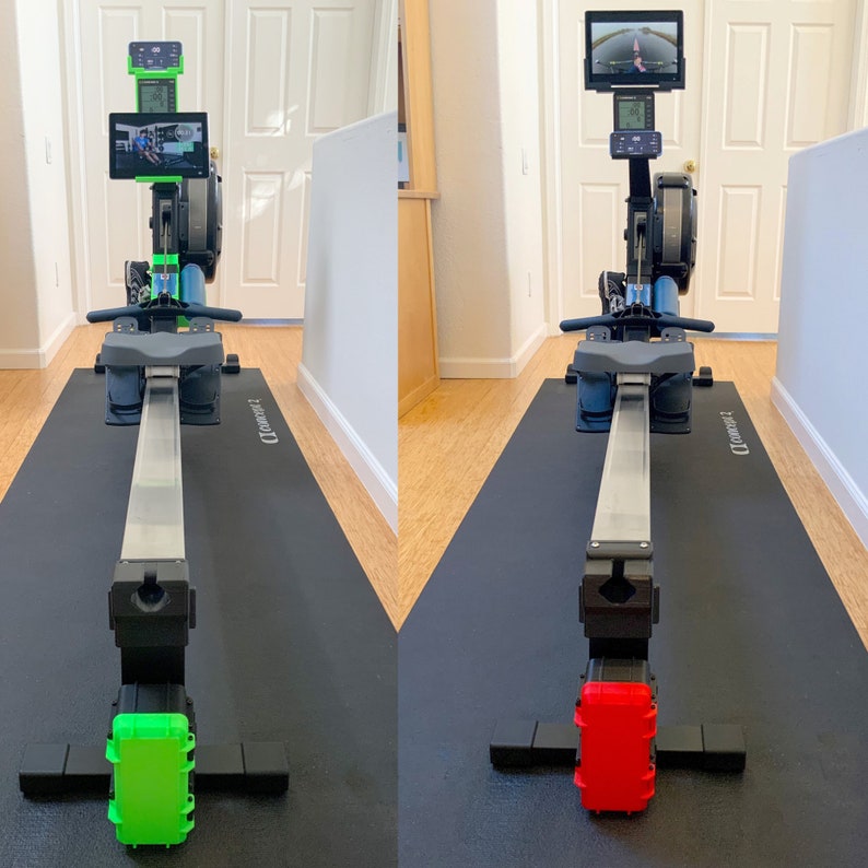 May include: A black Concept2 rowing machine with a digital display and a tablet holder. The machine is on a black mat. The image shows the machine with a green and a red protective case.