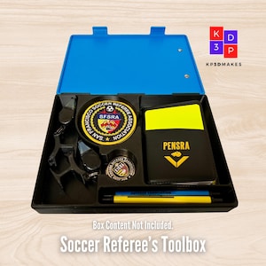 May include: A black plastic referee's toolbox with a blue lid. The toolbox is open and contains a whistle, a red and yellow card, a referee badge, and two yellow pens. The text "KP3DMAKES" is printed on the lid.